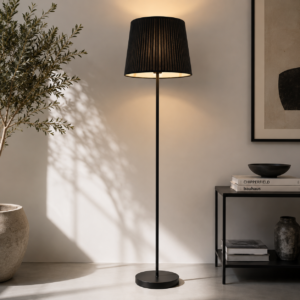 Lampada da terra nera con paralume plissettato illuminata, posta accanto a una pianta in vaso e un tavolino nero con libri.