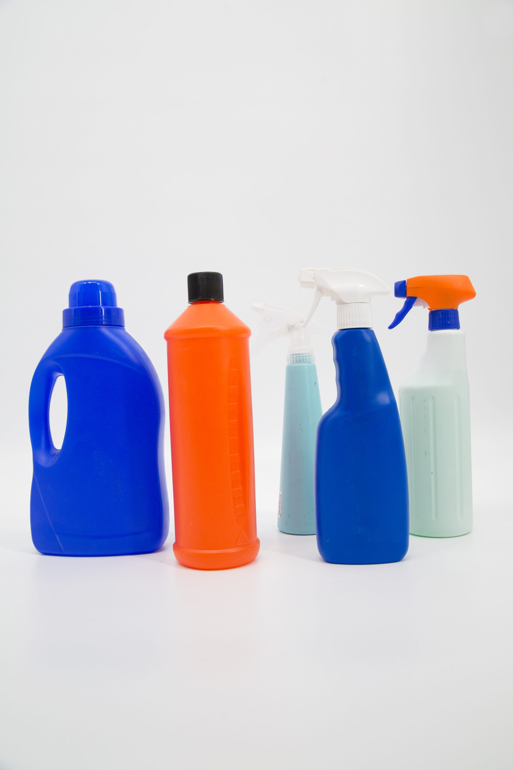 Cinque flaconi e spruzzini in plastica di vari colori (blu, arancione, azzurro, verde chiaro) disposti su sfondo bianco.