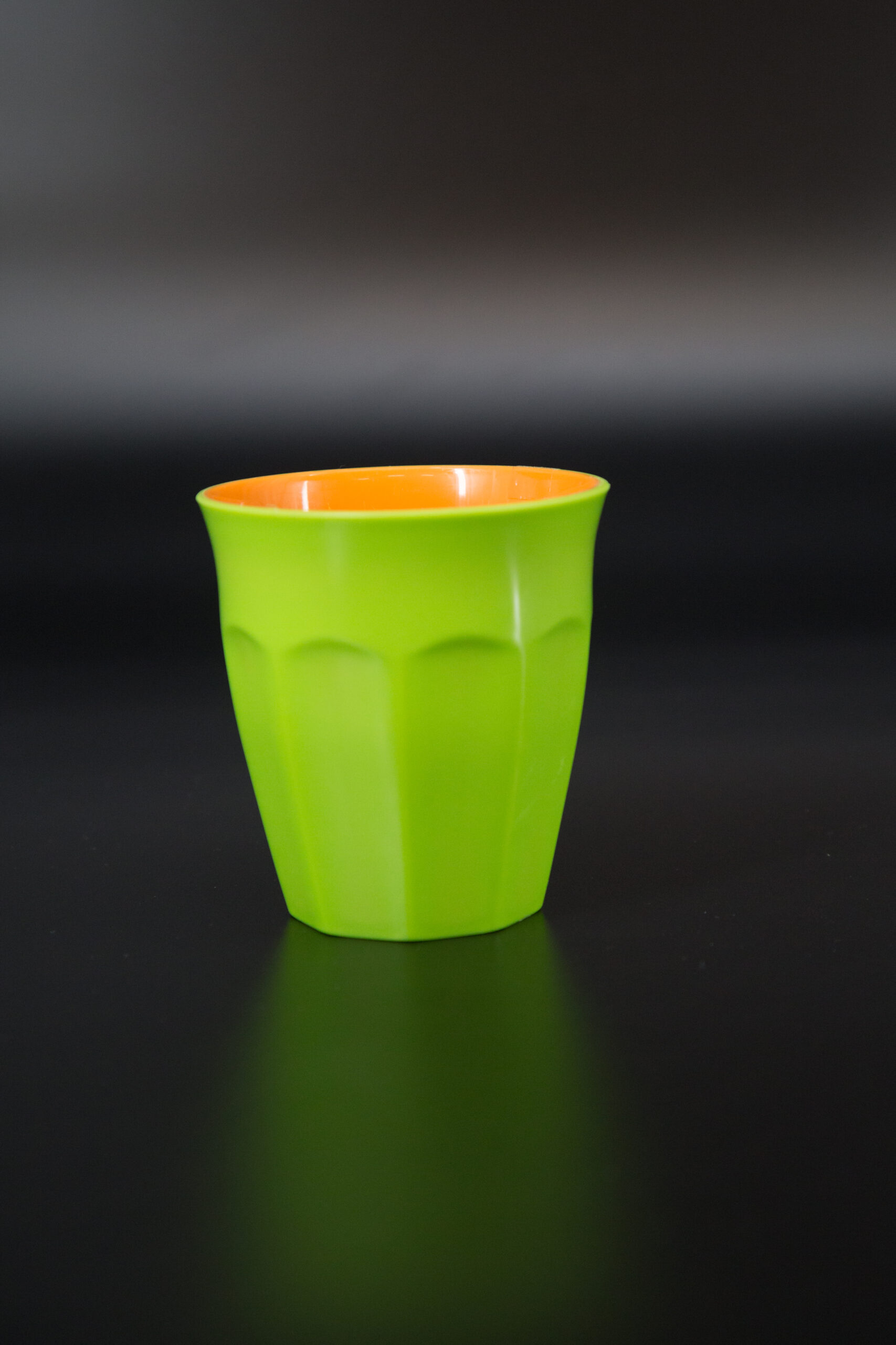Bicchiere in plastica bicolore verde acido all'esterno e arancione all'interno, con superficie facettata, su sfondo scuro.