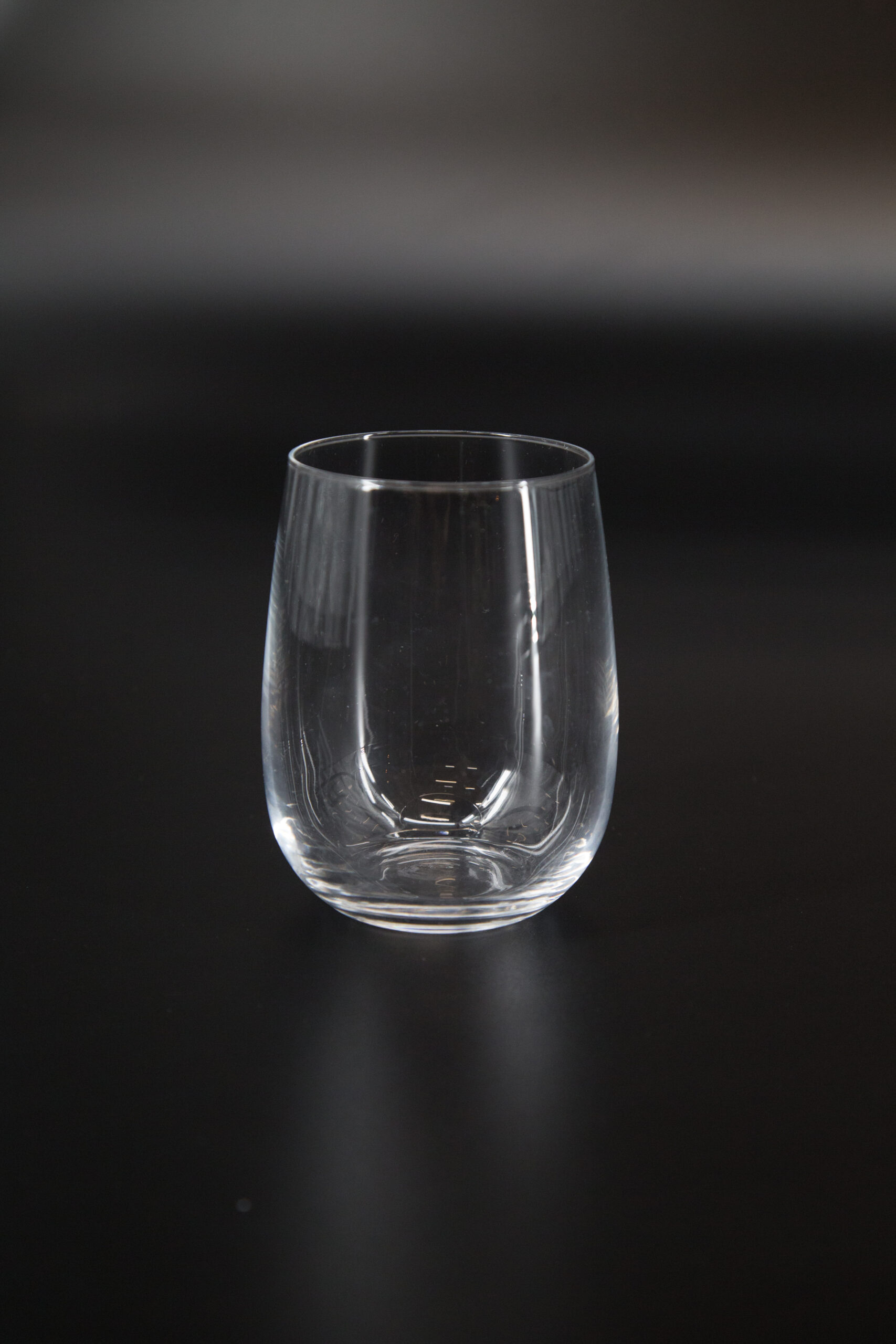 Bicchiere tumbler trasparente senza stelo, vuoto, su sfondo scuro, con riflessi di luce sulla superficie del vetro.