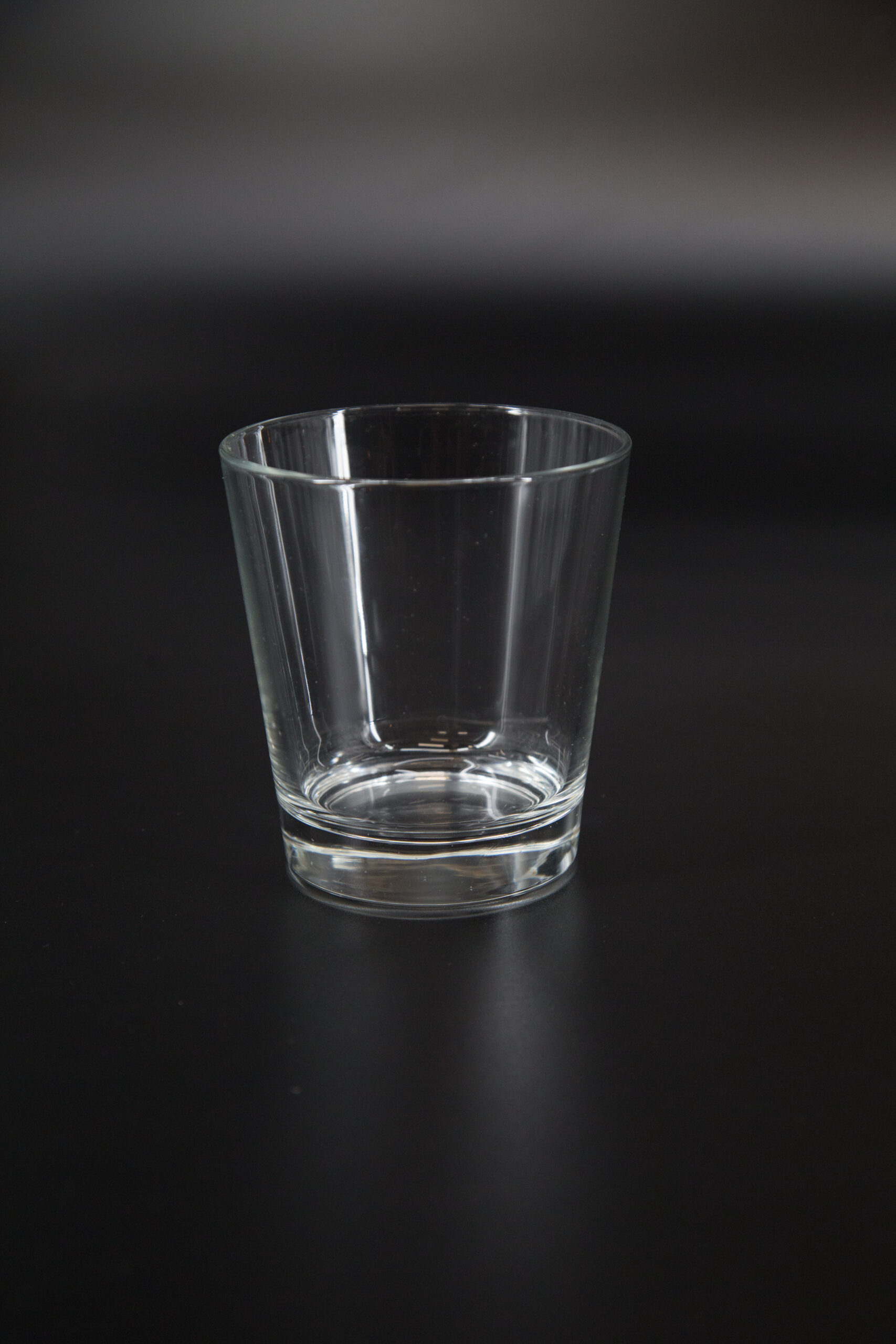 Un bicchiere tumbler basso in vetro trasparente, vuoto, posizionato su uno sfondo scuro e uniforme.