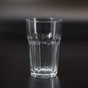 Bicchiere tumbler in vetro trasparente con scanalature verticali e base ottagonale, fotografato su sfondo scuro.