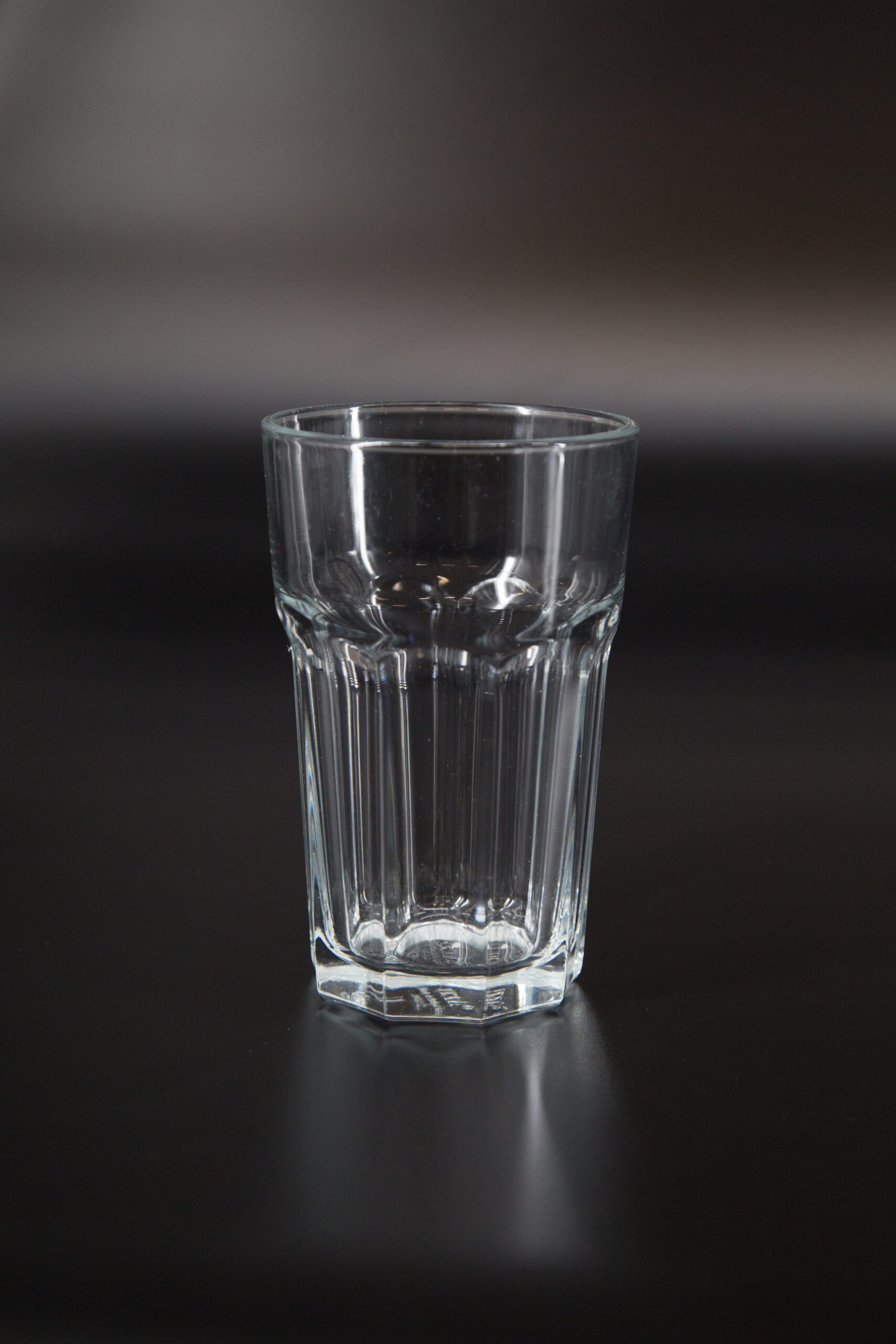 Bicchiere tumbler in vetro trasparente con scanalature verticali e base ottagonale, fotografato su sfondo scuro.