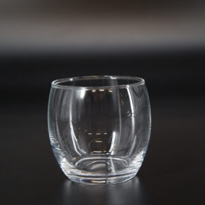Bicchiere tumbler basso in vetro trasparente, senza stelo, con base solida e corpo leggermente bombato, su sfondo scuro.