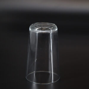 Bicchiere in vetro trasparente, di forma cilindrica conica, capovolto su sfondo scuro, con riflessi di luce sulla superficie.