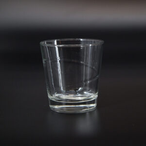 Bicchiere tumbler in vetro trasparente con design semplice e base spessa, fotografato su sfondo scuro.
