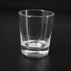 Bicchiere tumbler in vetro trasparente con base spessa, fotografato su sfondo nero, riflettendo la luce ambientale.