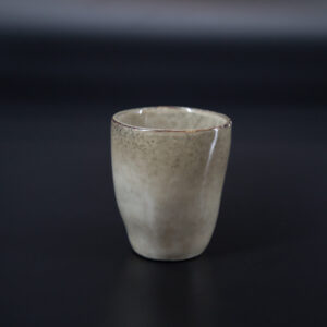 Bicchiere in ceramica artigianale di colore beige-grigio con finitura smaltata e bordo scuro, su sfondo nero.