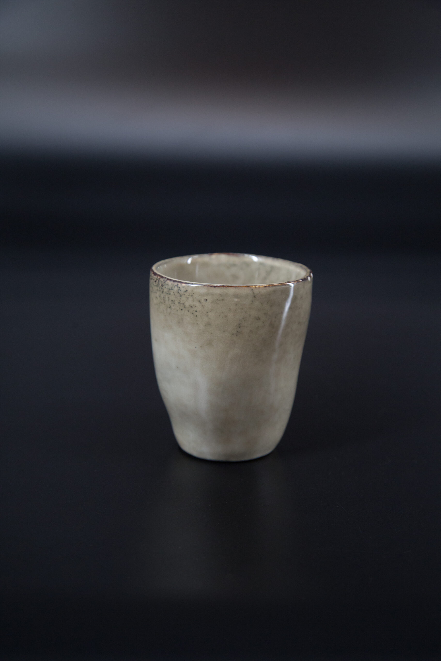 Bicchiere in ceramica artigianale di colore beige-grigio con finitura smaltata e bordo scuro, su sfondo nero.