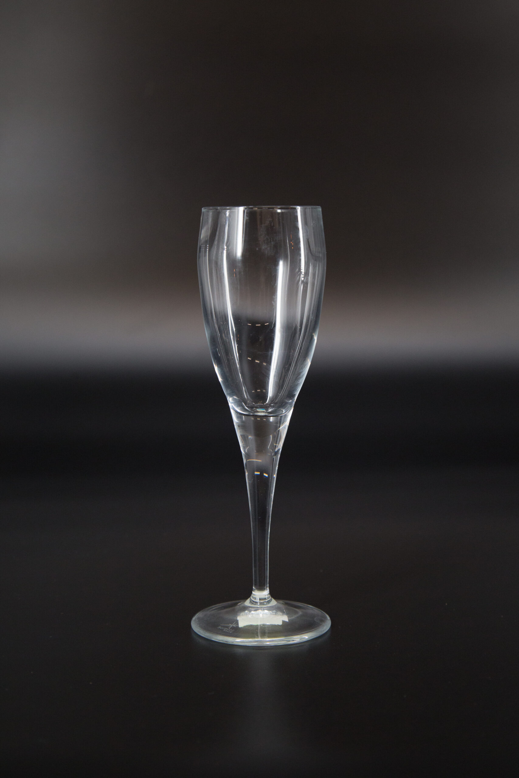 Calice flute in vetro trasparente con stelo sottile e base circolare, fotografato su sfondo scuro.