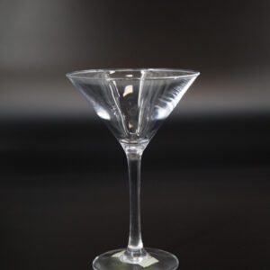 Un calice martini classico in vetro trasparente con stelo sottile e base circolare, fotografato su sfondo scuro.
