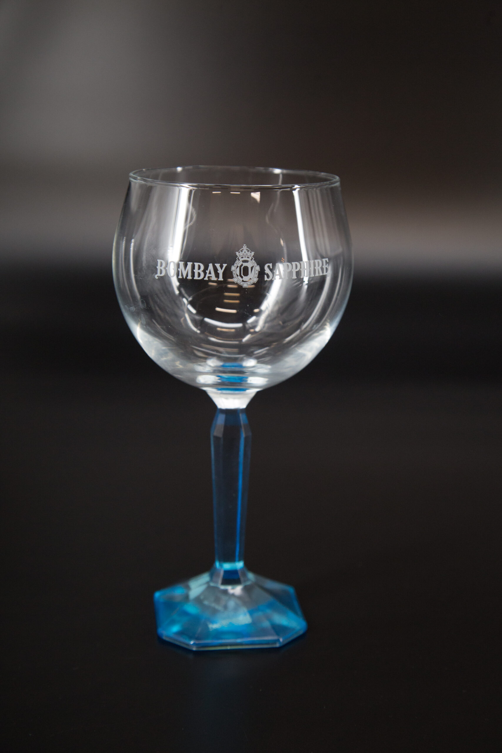 Calice da gin Bombay Sapphire in vetro trasparente con logo inciso, stelo e base blu sfaccettati, su sfondo scuro.