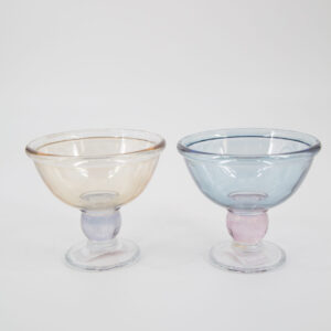Due coppette dessert in vetro con stelo colorato, una ambra con stelo viola e una azzurra con stelo rosa, su sfondo bianco.