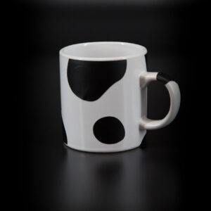 Tazza in ceramica bianca con grandi macchie nere irregolari, simile al manto di una mucca, su sfondo scuro.