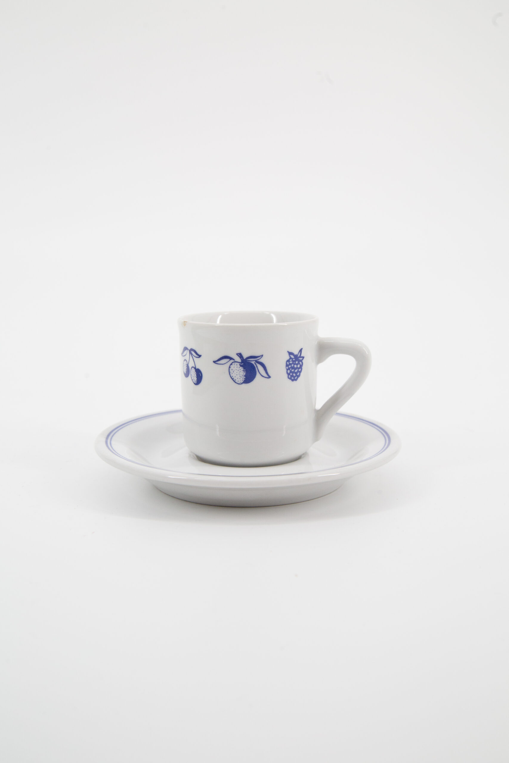 Set di tazza e piattino in ceramica bianca con decorazioni a frutta stilizzata blu e bordo blu, su sfondo bianco.