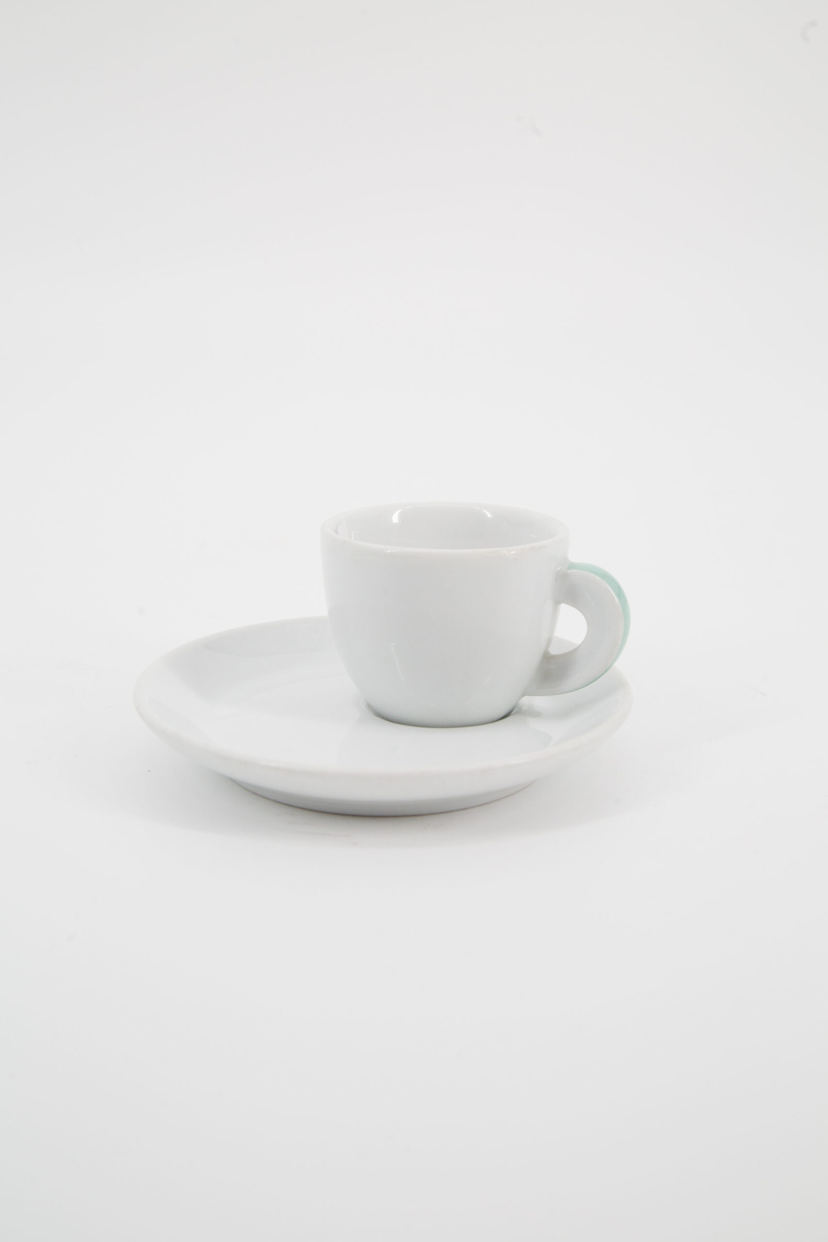 Set di tazzina e piattino espresso in ceramica bianca su sfondo neutro, con manico della tazzina leggermente verdastro.