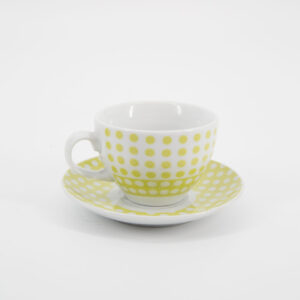 Tazza e piattino in ceramica bianca con motivo a pois gialli, posizionati centralmente su sfondo bianco.