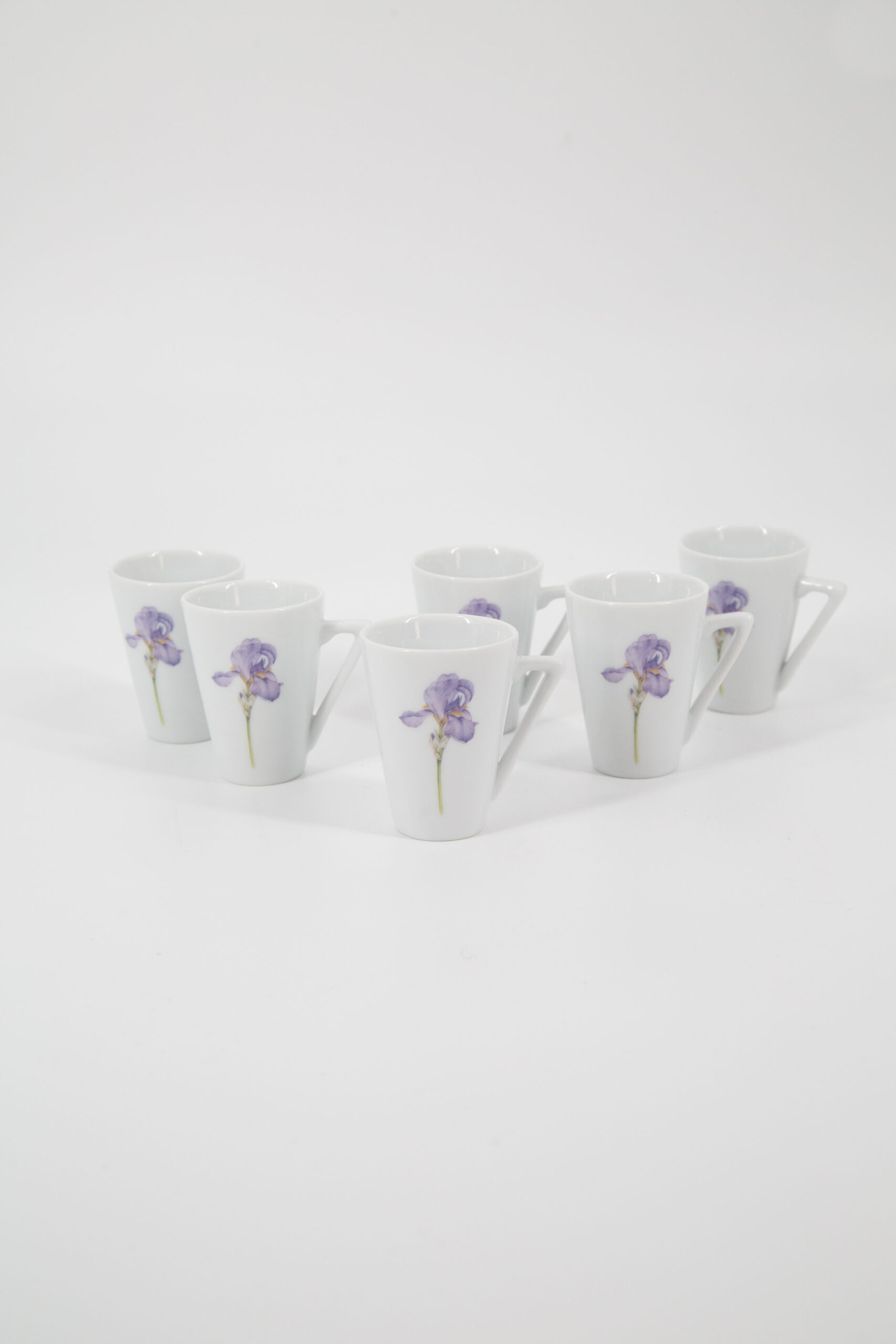 Sei tazzine da caffè in ceramica bianca con manico triangolare e decorazione di un fiore di iris viola, disposte su sfondo bianco.