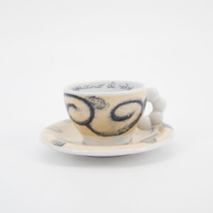 Set tazzina e piattino in ceramica color crema con decorazioni a spirale blu e manico a sfere, su sfondo bianco.