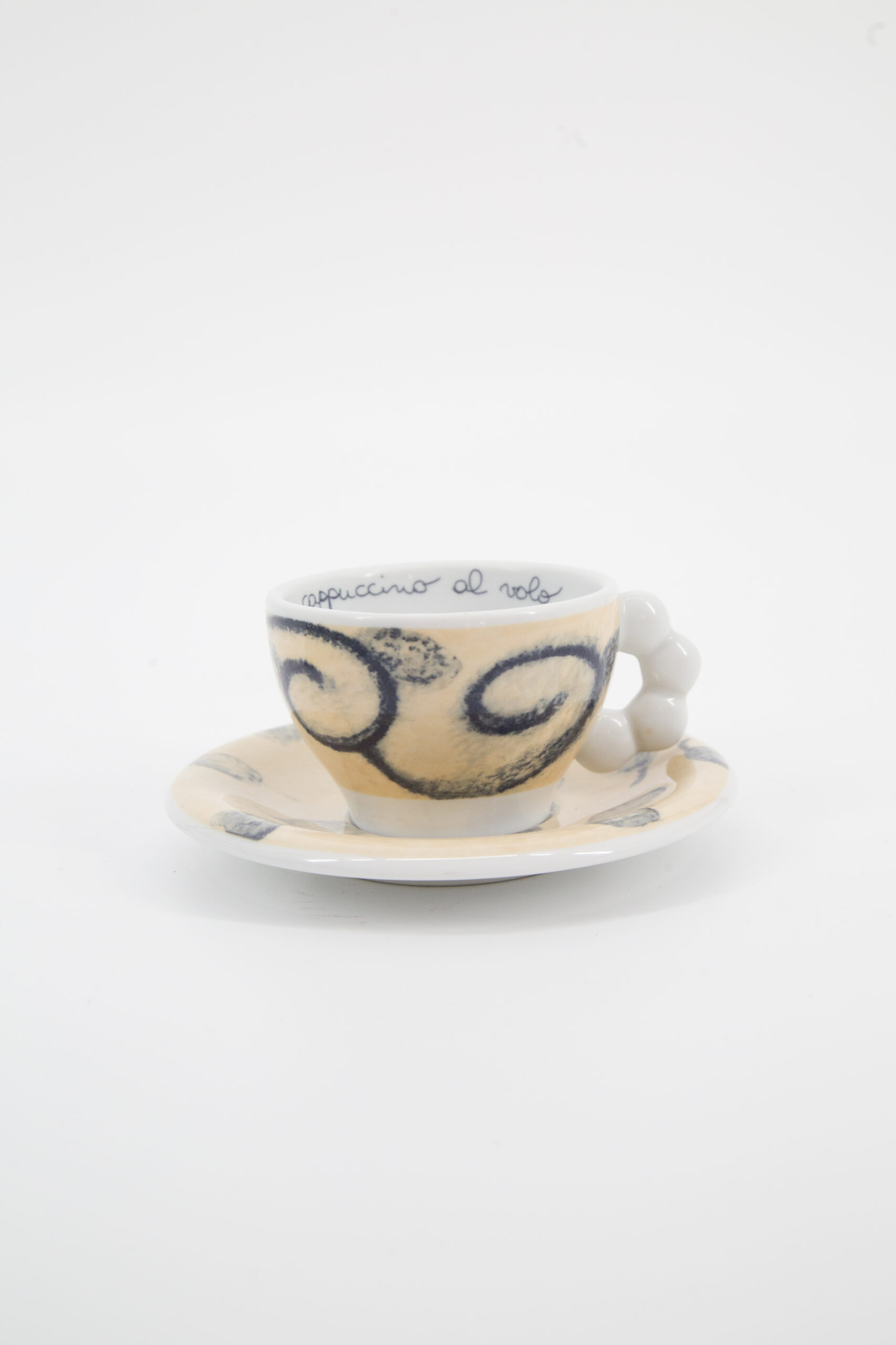 Set tazzina e piattino in ceramica color crema con decorazioni a spirale blu e manico a sfere, su sfondo bianco.