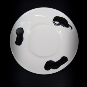Piatto in ceramica bianco con tre macchie nere irregolari, visto dall'alto su sfondo nero.