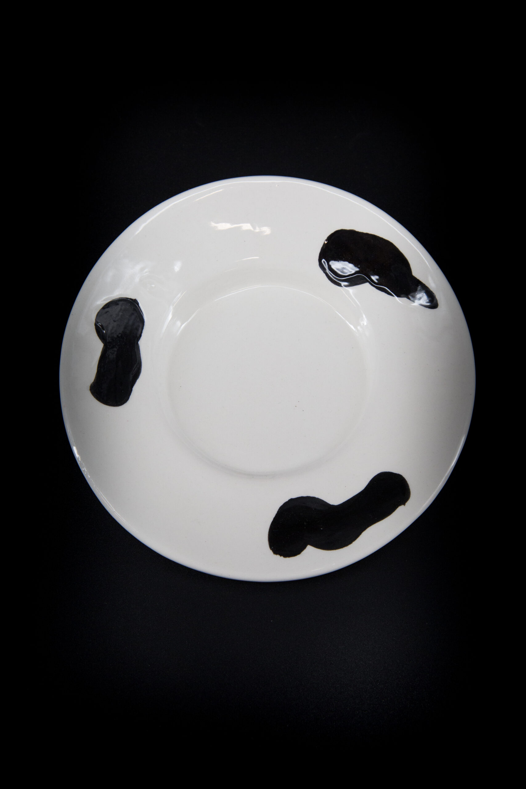 Piatto in ceramica bianco con tre macchie nere irregolari, visto dall'alto su sfondo nero.