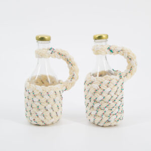 Due bottiglie in vetro trasparente con tappo dorato, rivestite in corda intrecciata ecrù con dettagli rossi e verdi, su sfondo bianco.