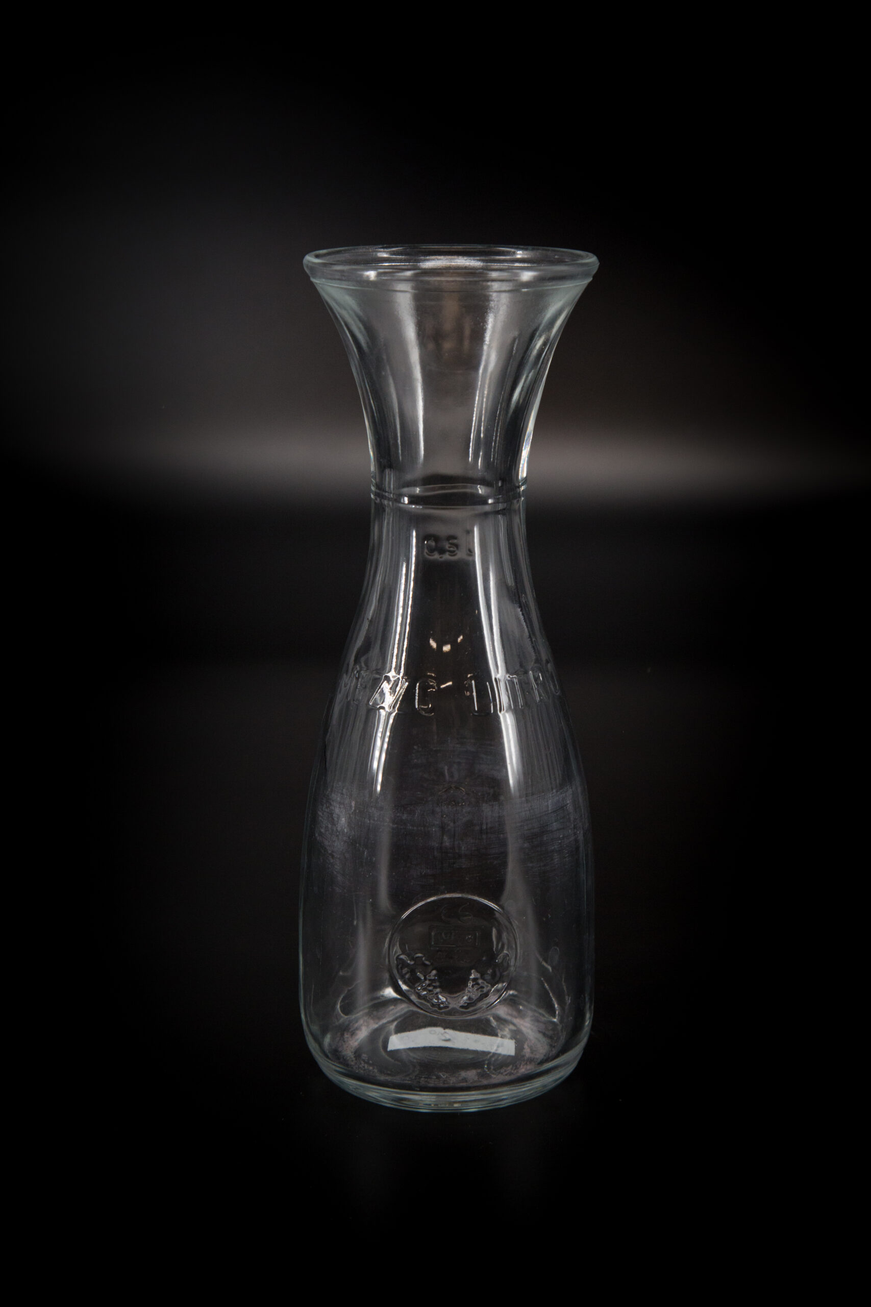 Caraffa in vetro trasparente da 0.5 litri con collo svasato e marcature impresse, fotografata su sfondo nero.
