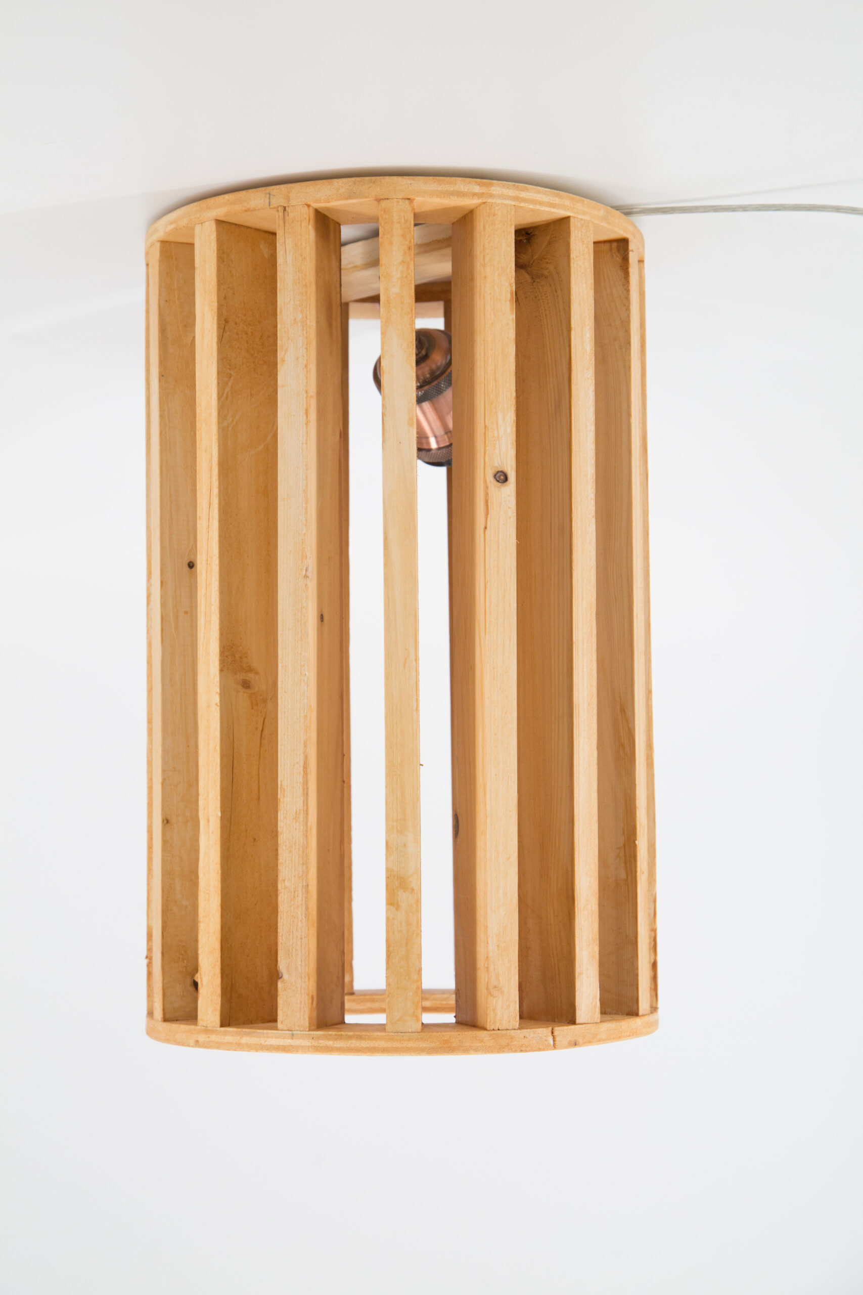 Paralume a sospensione cilindrico in legno chiaro a listelli verticali, con lampadina visibile all'interno, su sfondo bianco.