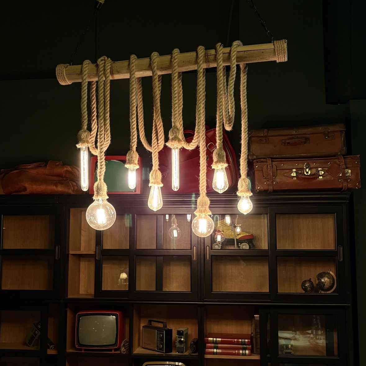 Lampadario a sospensione rustico con trave orizzontale in legno e otto lampadine a filamento stile Edison appese con corde spesse.