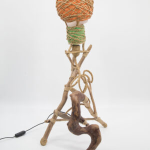 Lampada da tavolo artigianale con base in rami di legno naturale e paralume avvolto in corda di iuta e tessuto arancione e verde, su sfondo bianco.