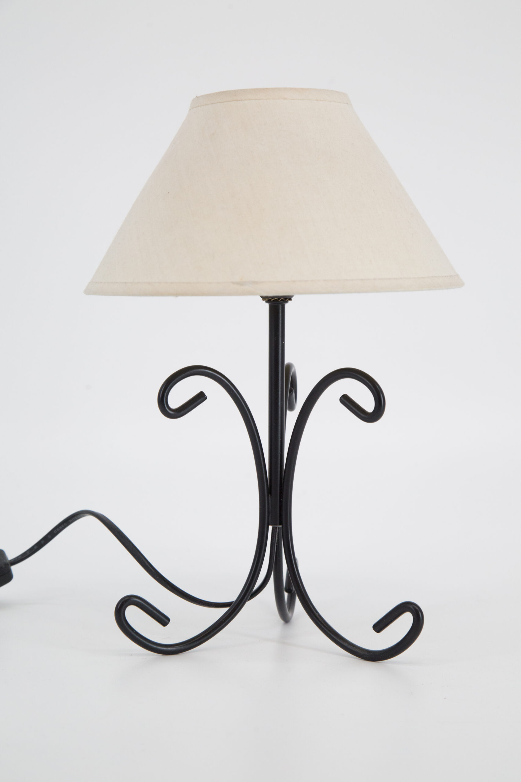 Lampada da tavolo con base in ferro battuto nero e paralume conico in tessuto ecrù, su sfondo bianco.