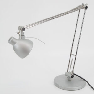 Lampada da scrivania articolata in metallo di colore argento su sfondo bianco, con braccio esteso e testa orientata.