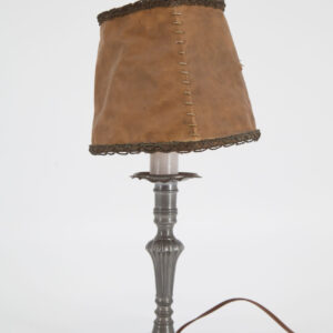 Lampada da tavolo con base in metallo grigio scuro e paralume in similpelle marrone chiaro con cuciture a vista e bordo decorato, su sfondo bianco.