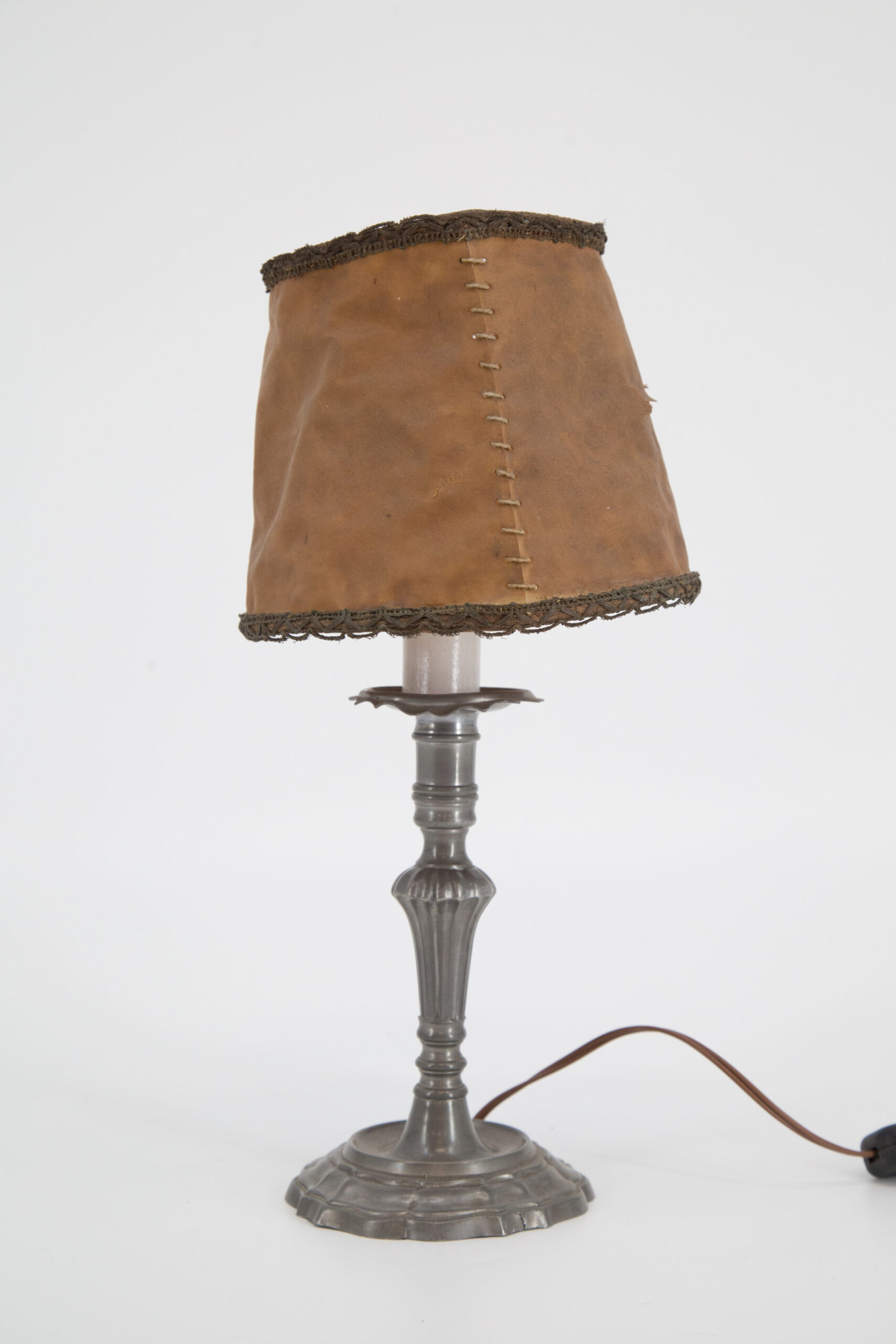 Lampada da tavolo con base in metallo grigio scuro e paralume in similpelle marrone chiaro con cuciture a vista e bordo decorato, su sfondo bianco.