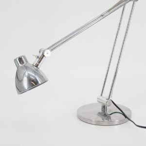 Lampada da scrivania cromata con braccio articolato e paralume a campana, su sfondo bianco. Ideale per noleggio in produzioni.