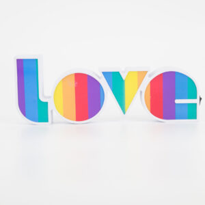 Insegna decorativa bianca con la parola "LOVE" riempita da strisce verticali arcobaleno, su sfondo bianco.