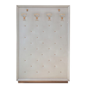 Pannello appendiabiti verticale grigio chiaro con texture, quattro ganci in ottone e borchie decorative, su sfondo bianco.