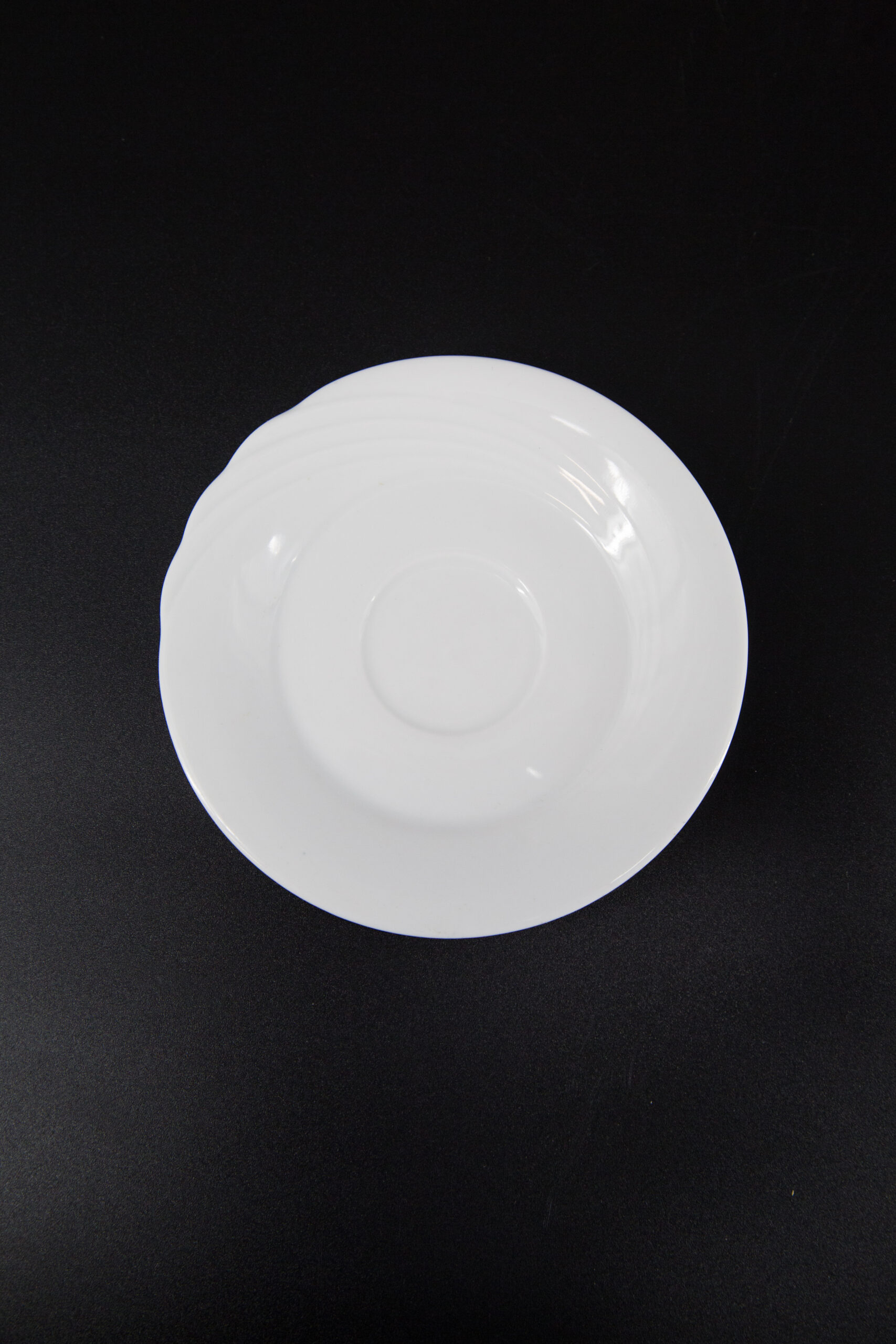 Sottotazza rotondo in ceramica bianca lucida, con bordo leggermente ondulato, posizionato su uno sfondo nero. Vista dall'alto.