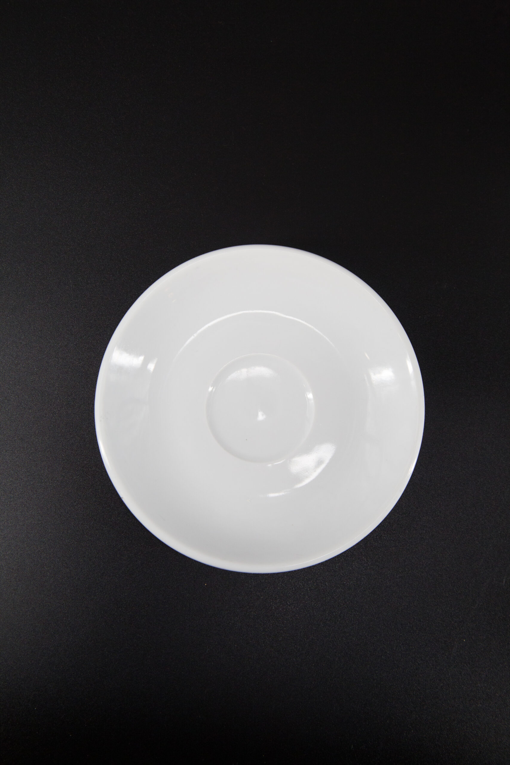 Un sottotazza bianco in ceramica, di forma circolare e con una leggera rientranza centrale, su sfondo nero.