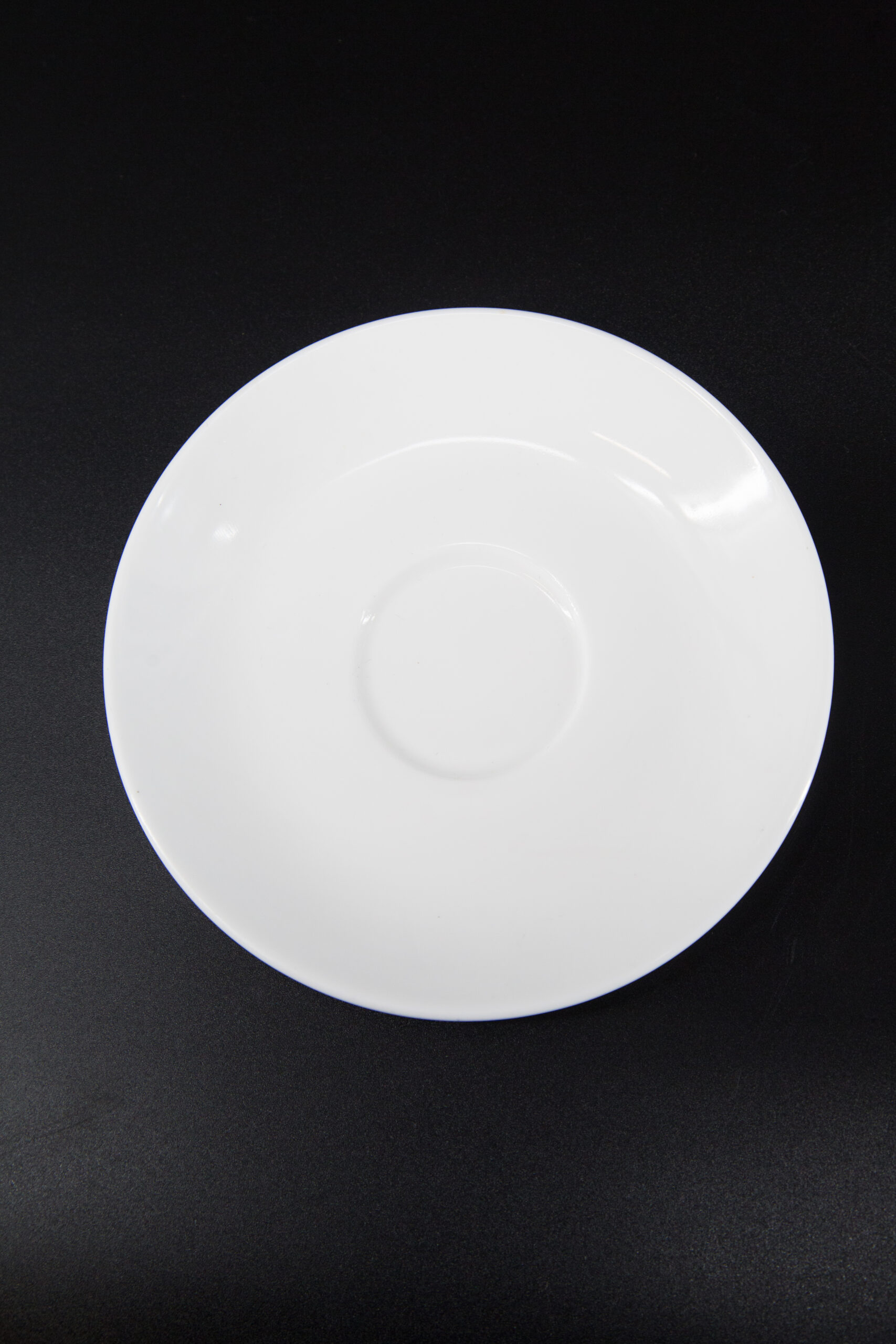 Piattino sottotazza bianco in ceramica o porcellana, visto dall'alto su sfondo nero, con incavo centrale per tazza.