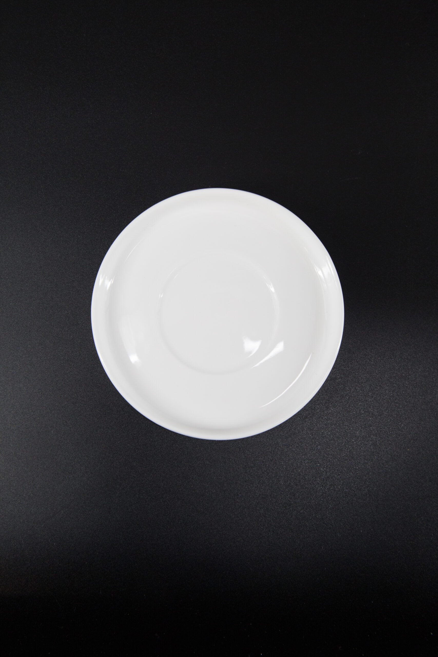 Un piattino bianco in ceramica, visto dall'alto, su uno sfondo nero, con una superficie liscia e lucida.