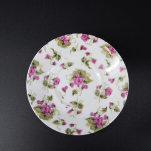 Piattino rotondo in ceramica bianca con un motivo floreale di fiori rosa e foglie verdi, su sfondo nero.