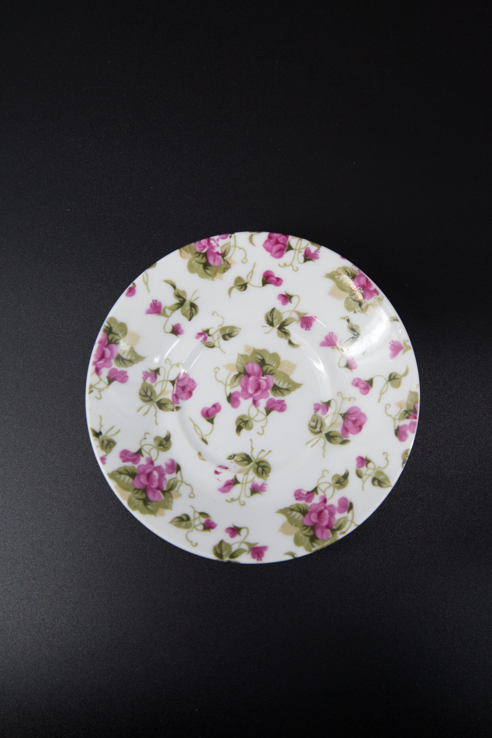 Piattino rotondo in ceramica bianca con un motivo floreale di fiori rosa e foglie verdi, su sfondo nero.