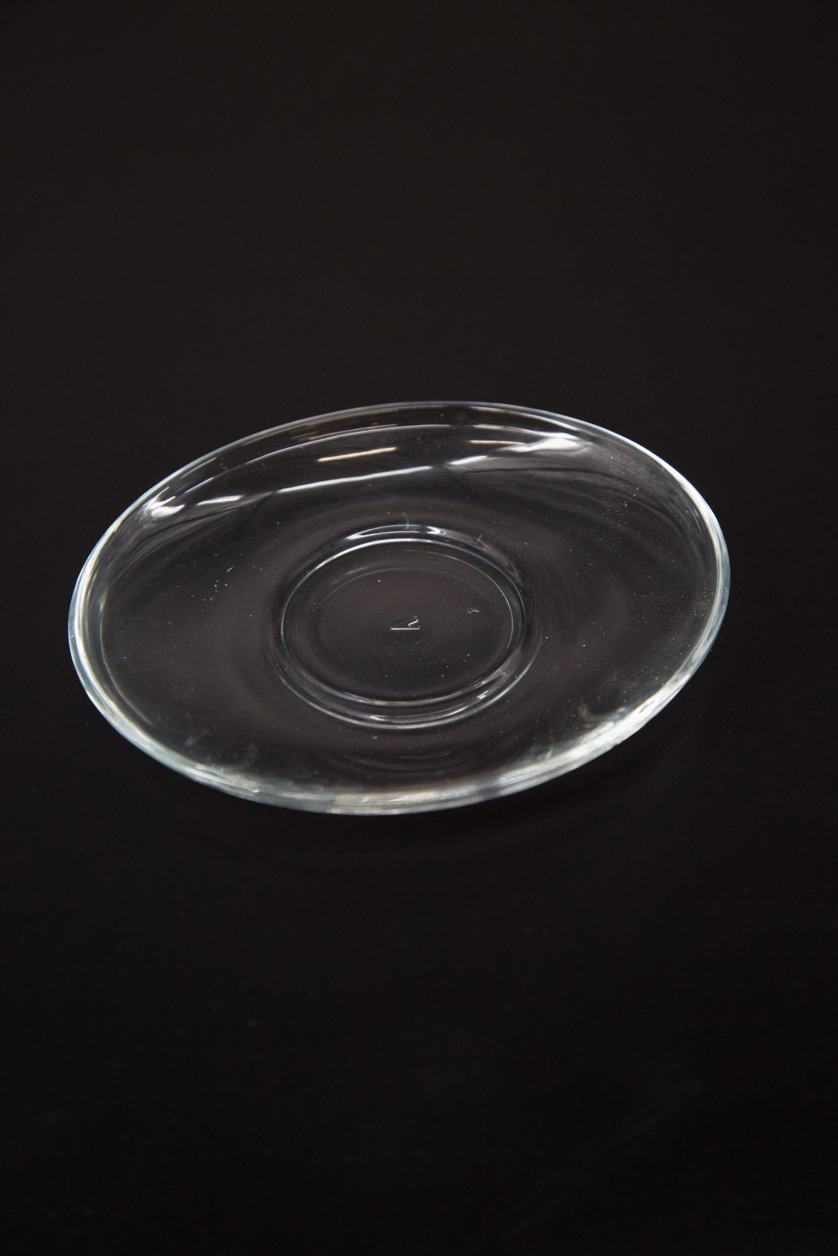 Piattino sottotazza in vetro trasparente, di forma circolare e design minimalista, posizionato su uno sfondo nero.