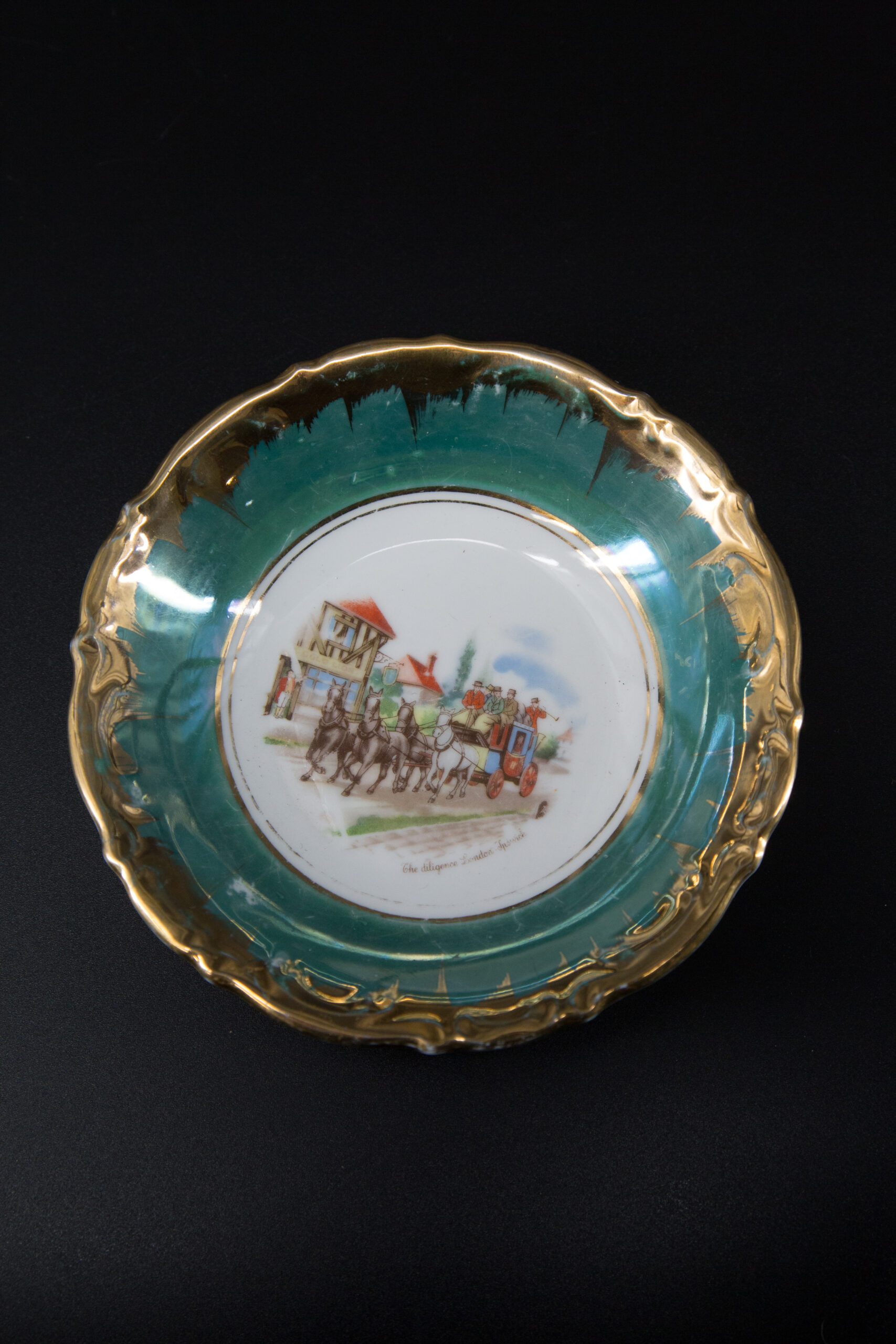 Piatto decorativo in ceramica con bordo dorato ondulato, fascia verde acqua e scena centrale di diligenza trainata da cavalli.