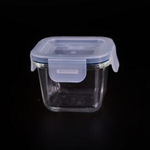 Contenitore quadrato in vetro trasparente con coperchio ermetico in plastica blu chiaro e quattro clip di chiusura, su sfondo nero.