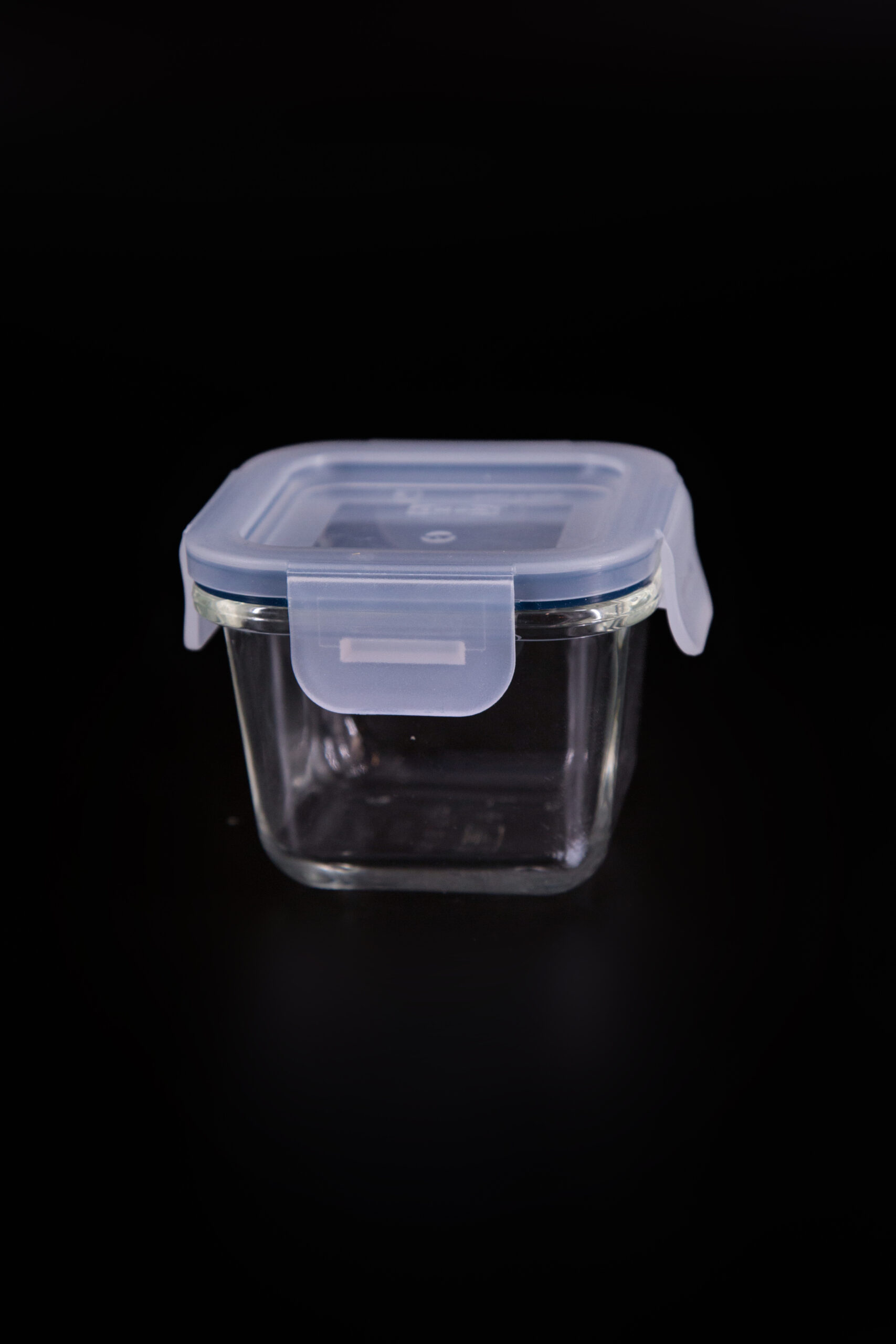 Contenitore quadrato in vetro trasparente con coperchio ermetico in plastica blu chiaro e quattro clip di chiusura, su sfondo nero.
