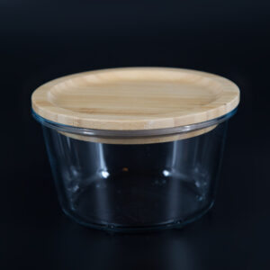 Contenitore cilindrico in vetro trasparente con coperchio piatto in legno di bambù chiaro, su sfondo scuro.