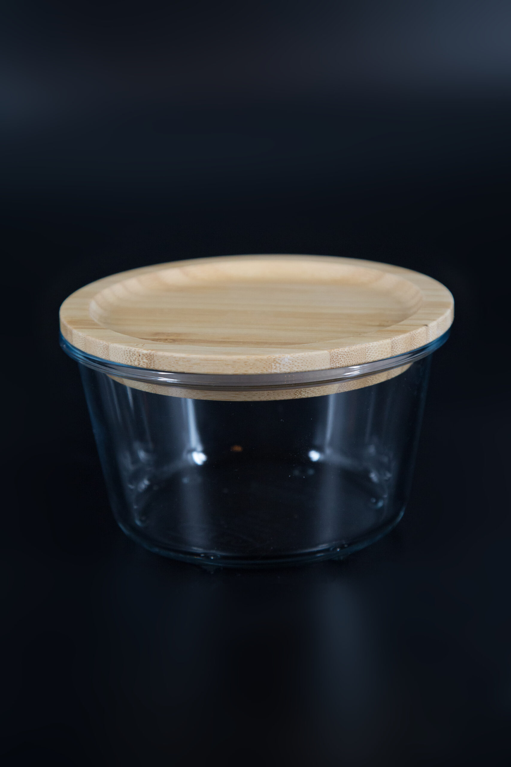 Contenitore cilindrico in vetro trasparente con coperchio piatto in legno di bambù chiaro, su sfondo scuro.